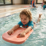 Young toddler boy in a blue and orange rash guard holding a pink foam kickboard while practicing kicking techniques in an indoor swimming pool.