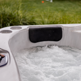 Tropic Seas Spas Lounge series charcoal gray headrest pillow installed in a white acrylic hot tub with bubbling water, featuring a grassy backyard lawn backdrop.
