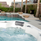 Tropic Seas Spas Lounge series charcoal gray headrest pillow installed in a white acrylic hot tub corner, set against a luxurious backyard swimming pool patio landscape.