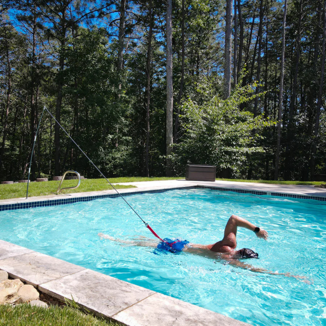 An athlete training in an outdoor inground pool using a Swim Tether stationary system, attached via a black resistance cord and red strap to a flexible vertical pole mounted on the grassy poolside edge, surrounded by green trees.