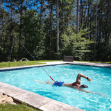 An athlete training in an outdoor inground pool using a Swim Tether stationary system, attached via a black resistance cord and red strap to a flexible vertical pole mounted on the grassy poolside edge, surrounded by green trees.
