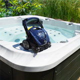 Outdoor lifestyle view of the Zodiac SPABOT™ SB1000 robotic spa cleaner resting on the white rim ledge of an above-ground hot tub spa, showcasing its dark blue chassis, large carrying handle, front scrubbing brushes, grey side wheels, and clear spa water in the background.